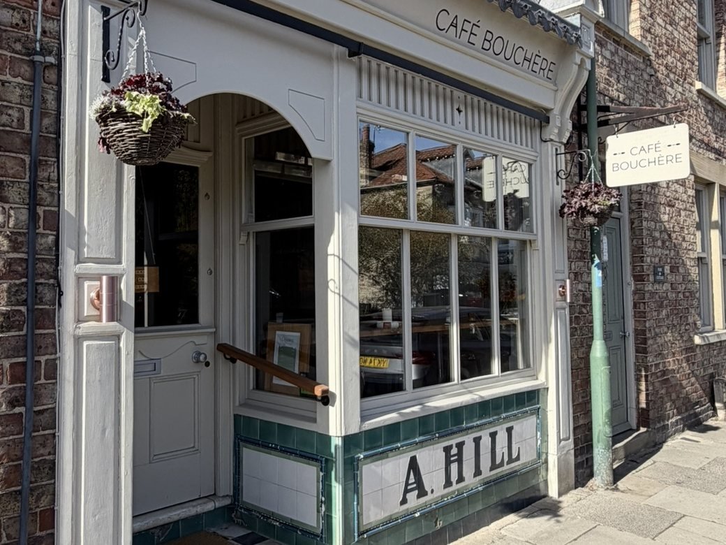 The Café Bouchère shopfront on Maltongate, Thornton-le-Dale, with a cream-painted bay window and arched doorway beneath the CAFÉ BOUCHÈRE fascia, a hanging sign to the right, and the name A. HILL in green-bordered tiles below the window