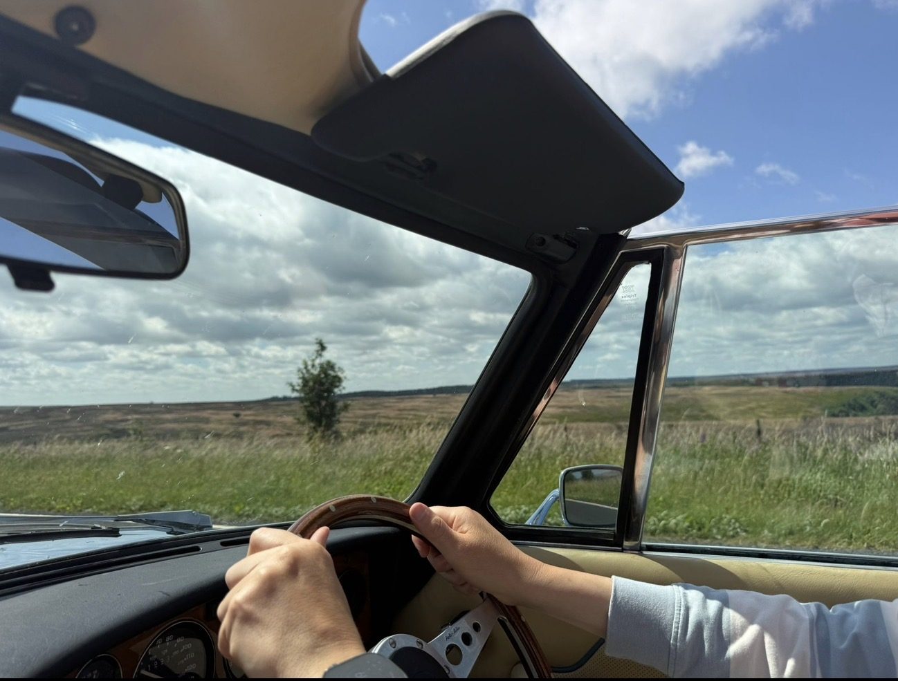 View from inside a moving Triumph Stag with the roof down: hands on a wood-rimmed steering wheel, the instrument dials ahead and open moorland through the windscreen.