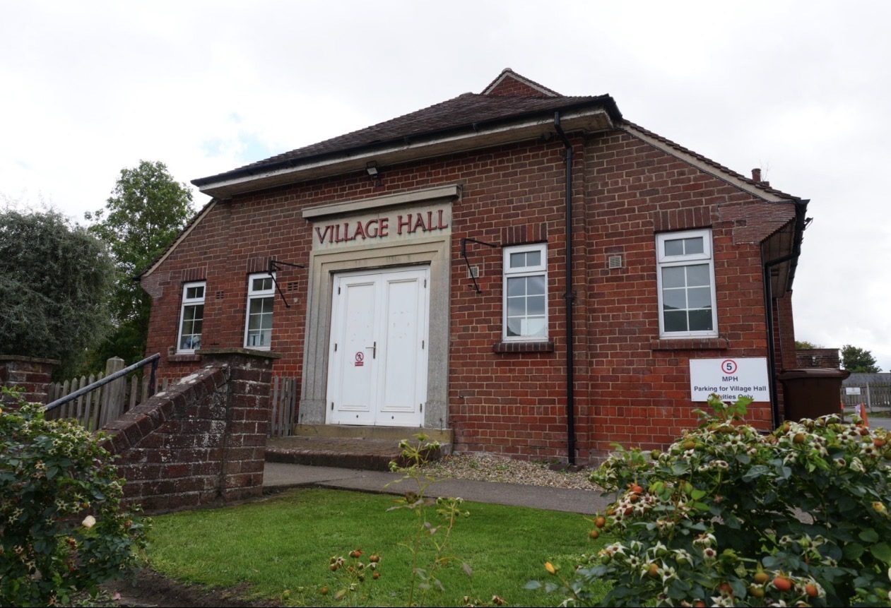 Thornton-le-Dale Village Hall on Pickering Road: a red-brick hall with a hipped tiled roof, stone trim around white double doors, and the words Village Hall in red letters above the entrance