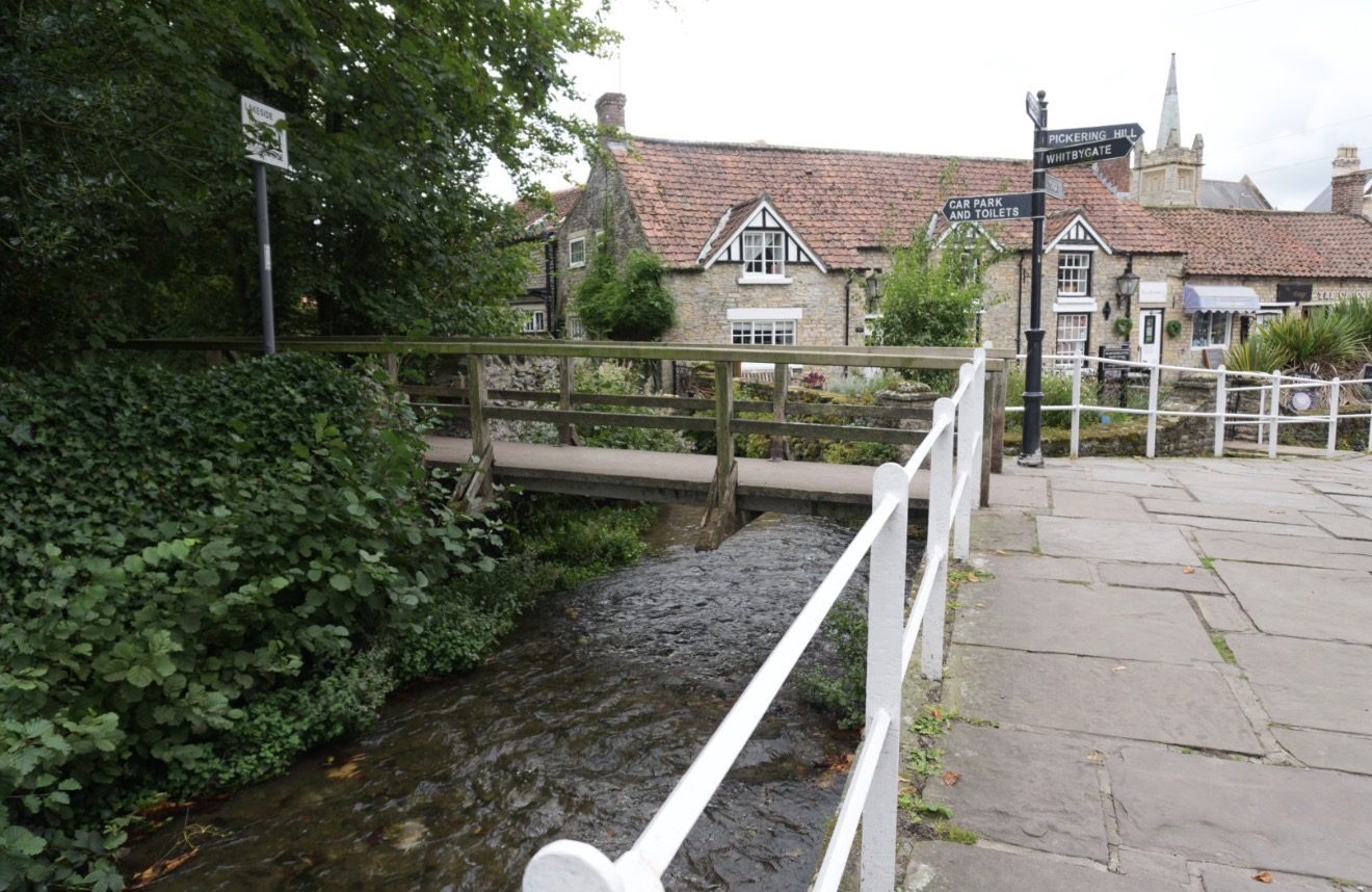 A wooden footbridge over Thornton Beck beside a fingerpost signed Pickering Hill, Whitbygate, and car park and toilets, with stone cottages and the church tower behind