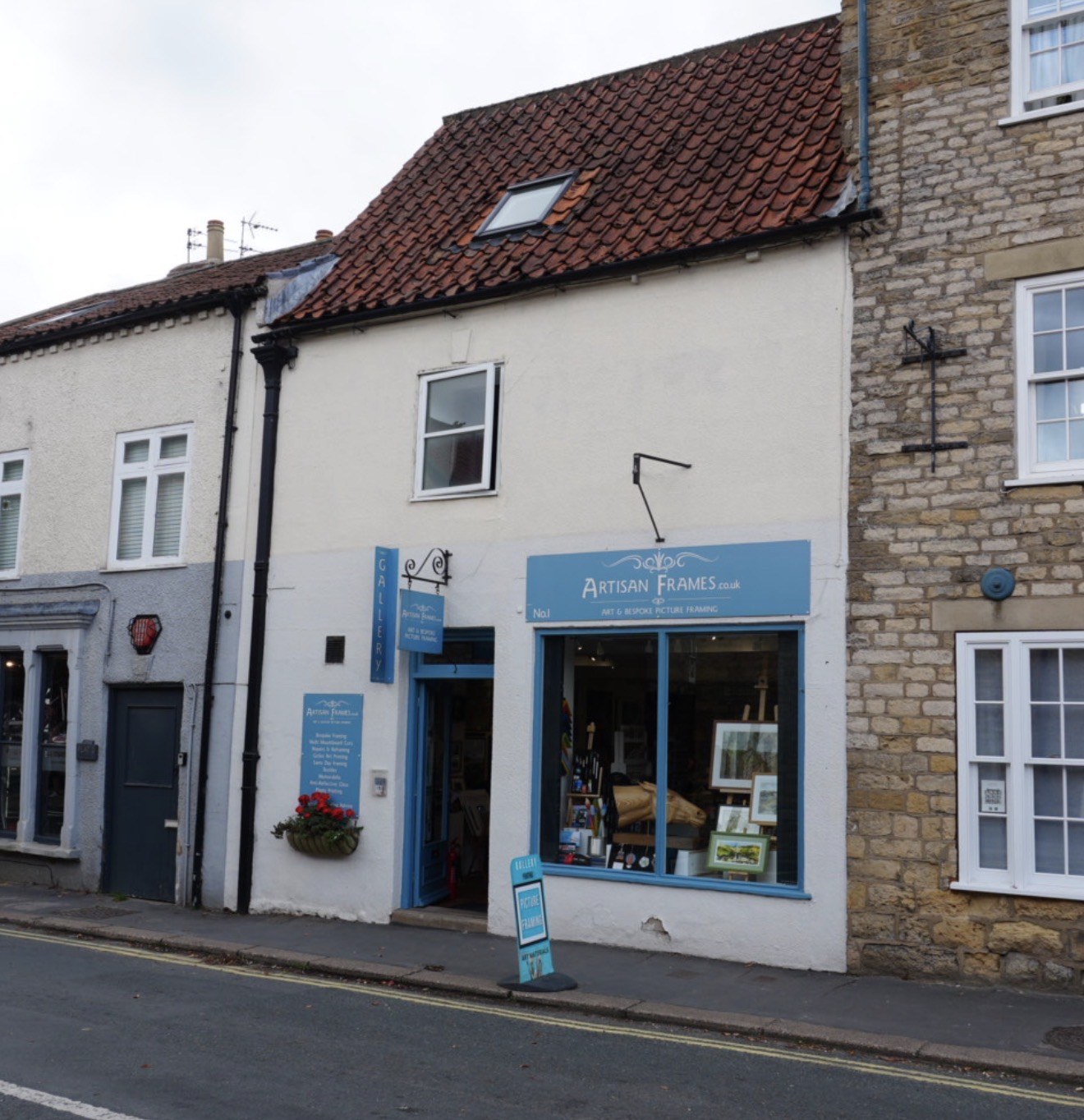The Artisan Frames studio and gallery shopfront at 1 Whitbygate in Thornton-le-Dale: a white-painted corner building with a blue sign reading Artisan Frames, a hanging gallery sign and framed artwork in the window