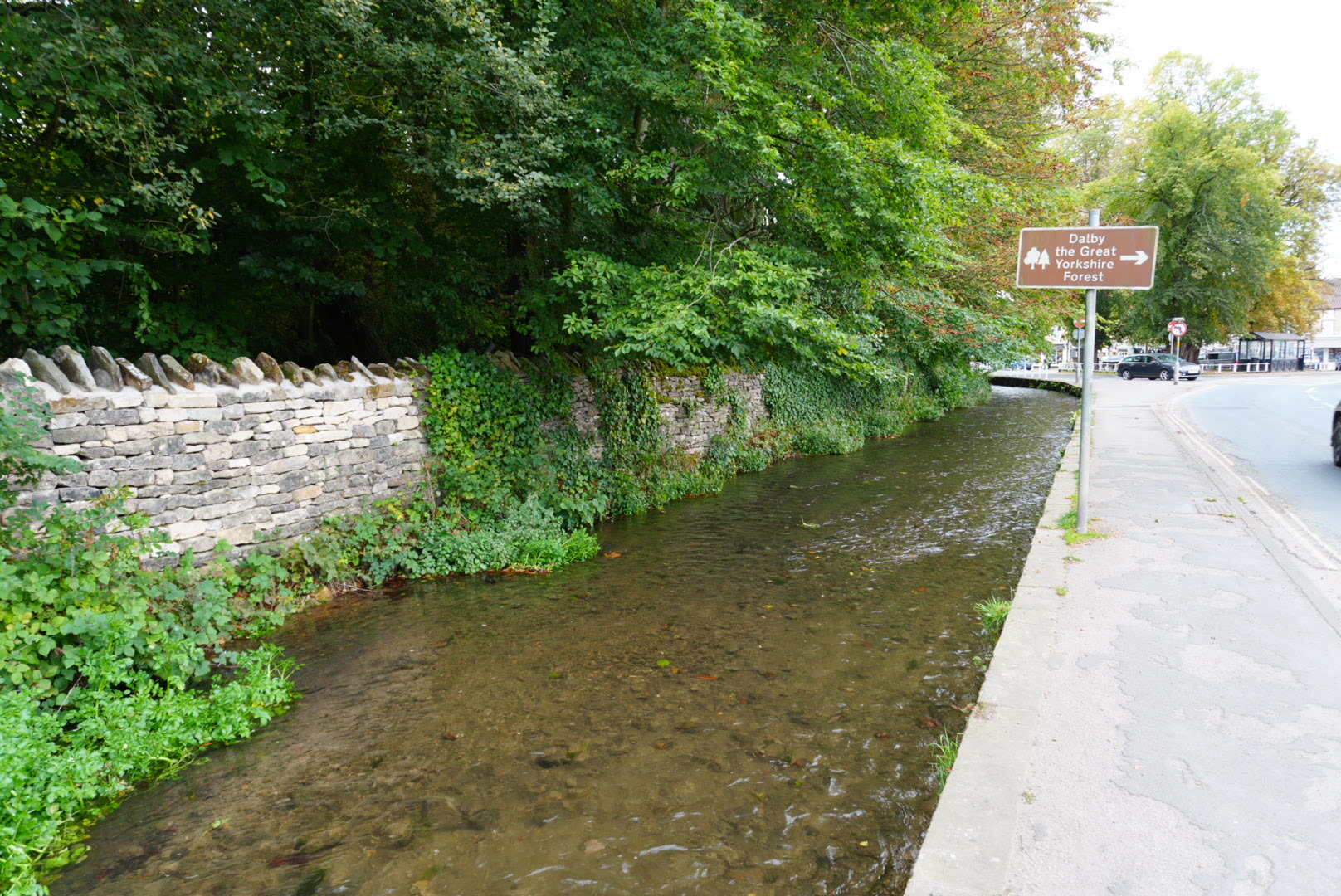 Thornton Beck beside the pavement at the edge of Thornton-le-Dale, with the brown tourist sign for Dalby Forest on the corner