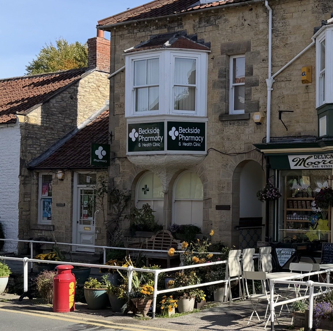 The stone frontage of Beckside Pharmacy and Health Clinic on Maltongate in Thornton-le-Dale, with a green pharmacy sign bearing a white cross, a first-floor bay window and Moore's delicatessen alongside
