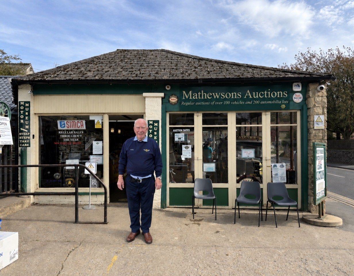 Bubbles, in a navy jumper and trousers, standing outside the Mathewsons Auctions building in Thornton-le-Dale, with its green fascia, stone-tiled roof, motor museum signs and a row of grey chairs beneath the windows