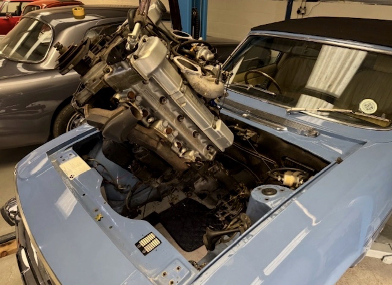 A Triumph V8 engine on a hoist strap being lowered into the engine bay of a light blue Triumph Stag in a workshop, with the bonnet off and other classic cars parked alongside.