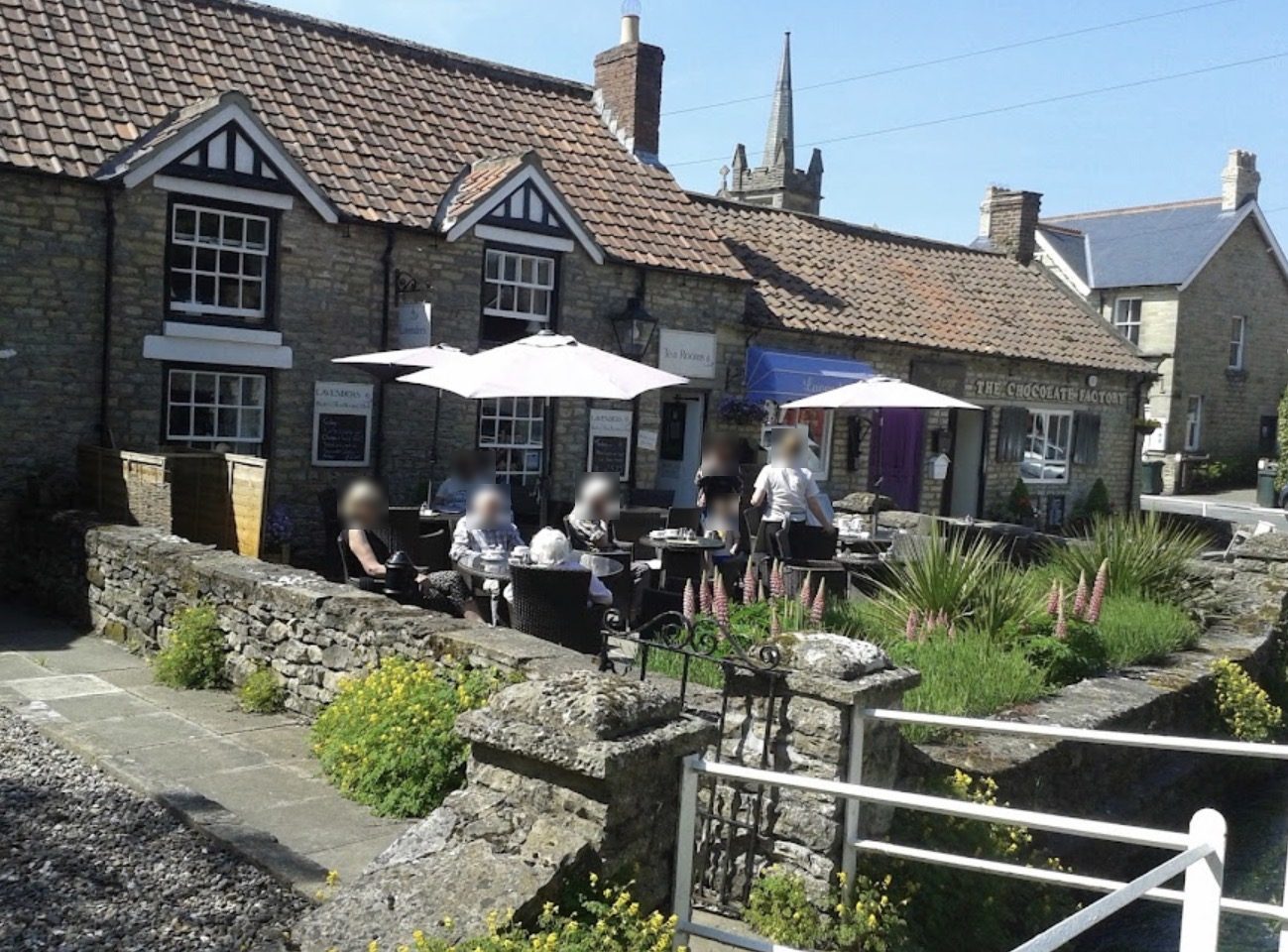 People at tables under parasols in a tea garden in front of stone cottages, with a church spire behind