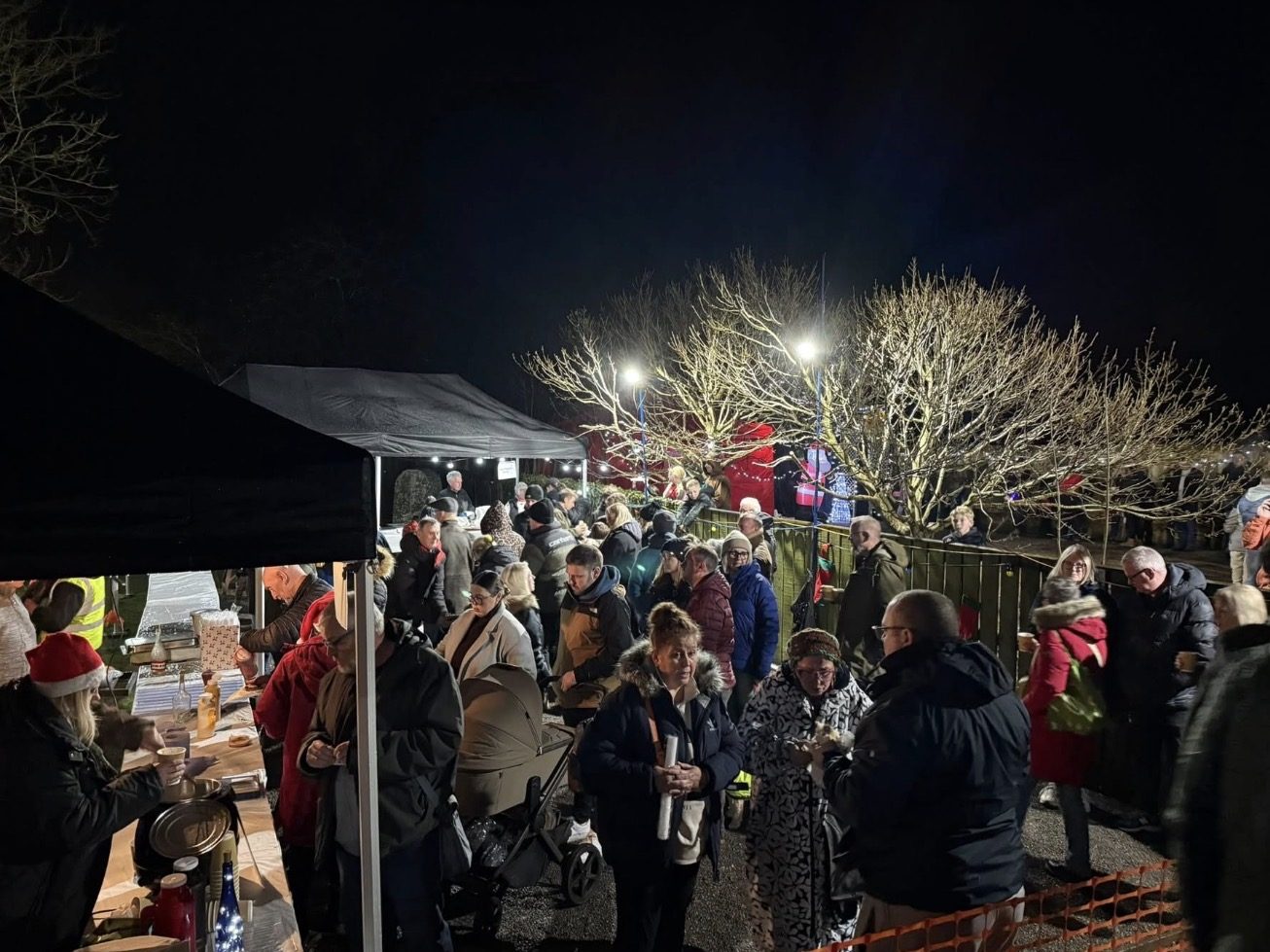 People gather at lit Christmas stalls beside bare trees and railings in Thornton-le-Dale at night.
