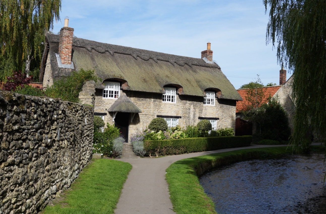The thatched Beck Isle Cottage seen across Thornton Beck, with a weeping willow on the right and a stone wall on the left