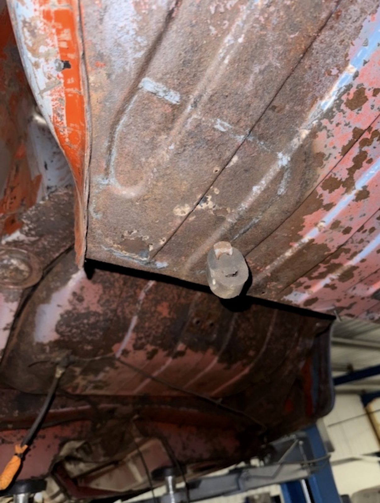 The rusted underside floorpan of a Triumph Stag seen from below on a workshop lift, with stamped reinforcement ribs, surface rust and patches of red primer.