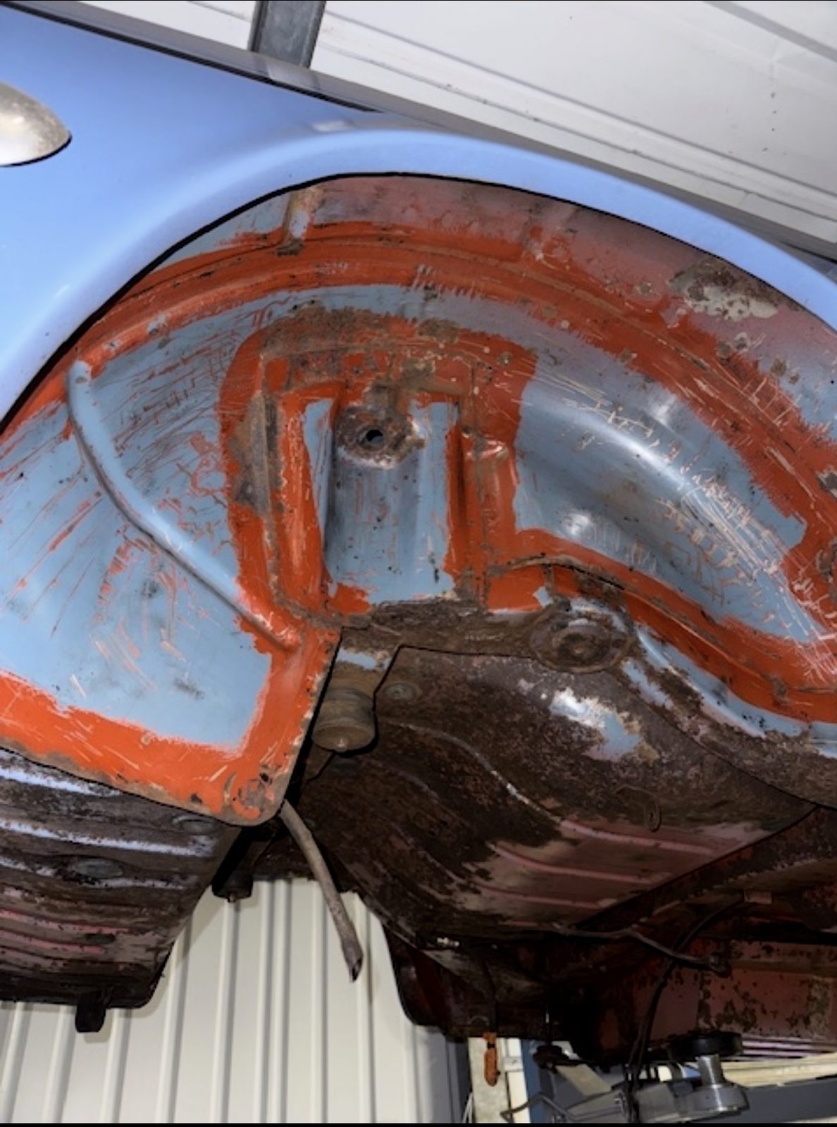 The underside of a Triumph Stag's wheel arch on a lift, with light blue outer paint, red primer along the seams and repaired areas, and patches of surface rust.