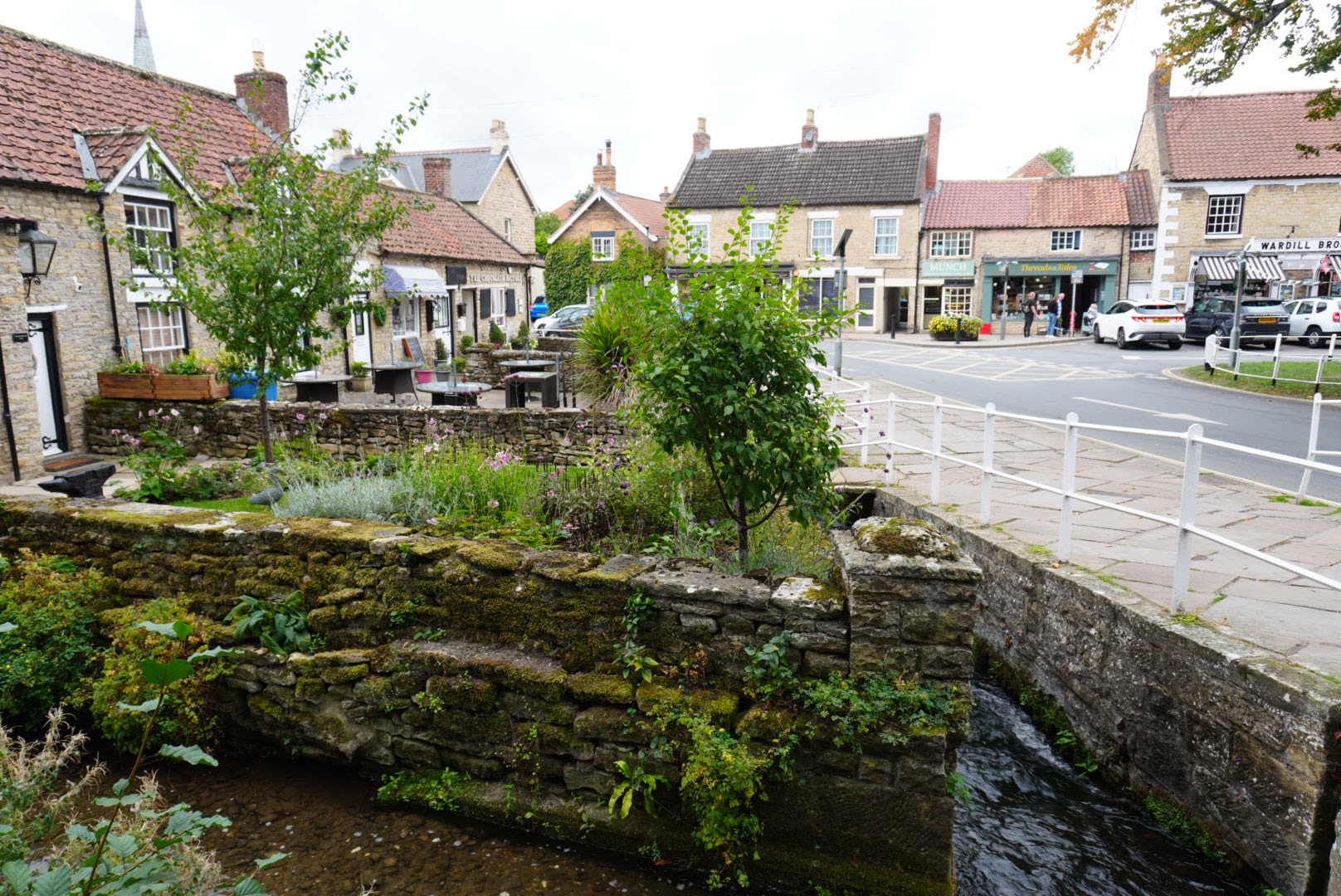Thornton Beck passing beneath a stone bridge at the corner of the village green, with gardens and village shops beyond