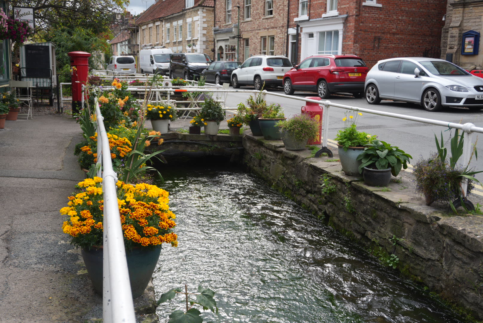 Flower pots of marigolds and dahlias lining the stone channel of Thornton Beck, with a red postbox, white railings and the shops of the main street behind
