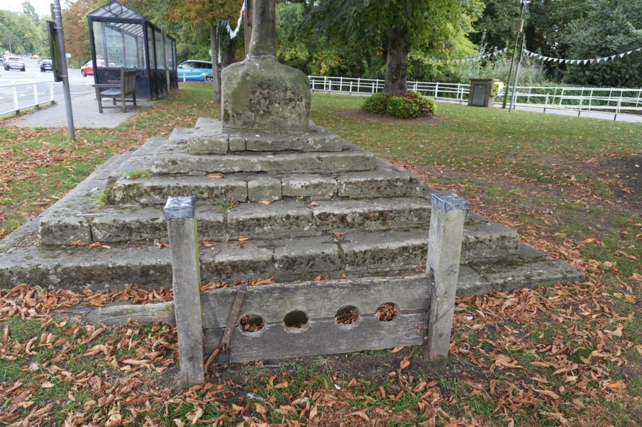 The stepped stone base of the market cross on the green, with the wooden village stocks in front among fallen leaves