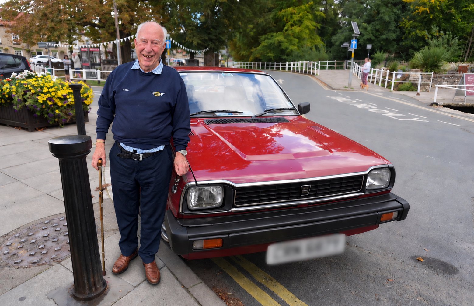 Bubbles, smiling, in a navy jumper embroidered with his name, leaning on the bonnet of a red hatchback parked by the beck in the centre of Thornton-le-Dale, with flower tubs, bunting and the white beckside railings behind
