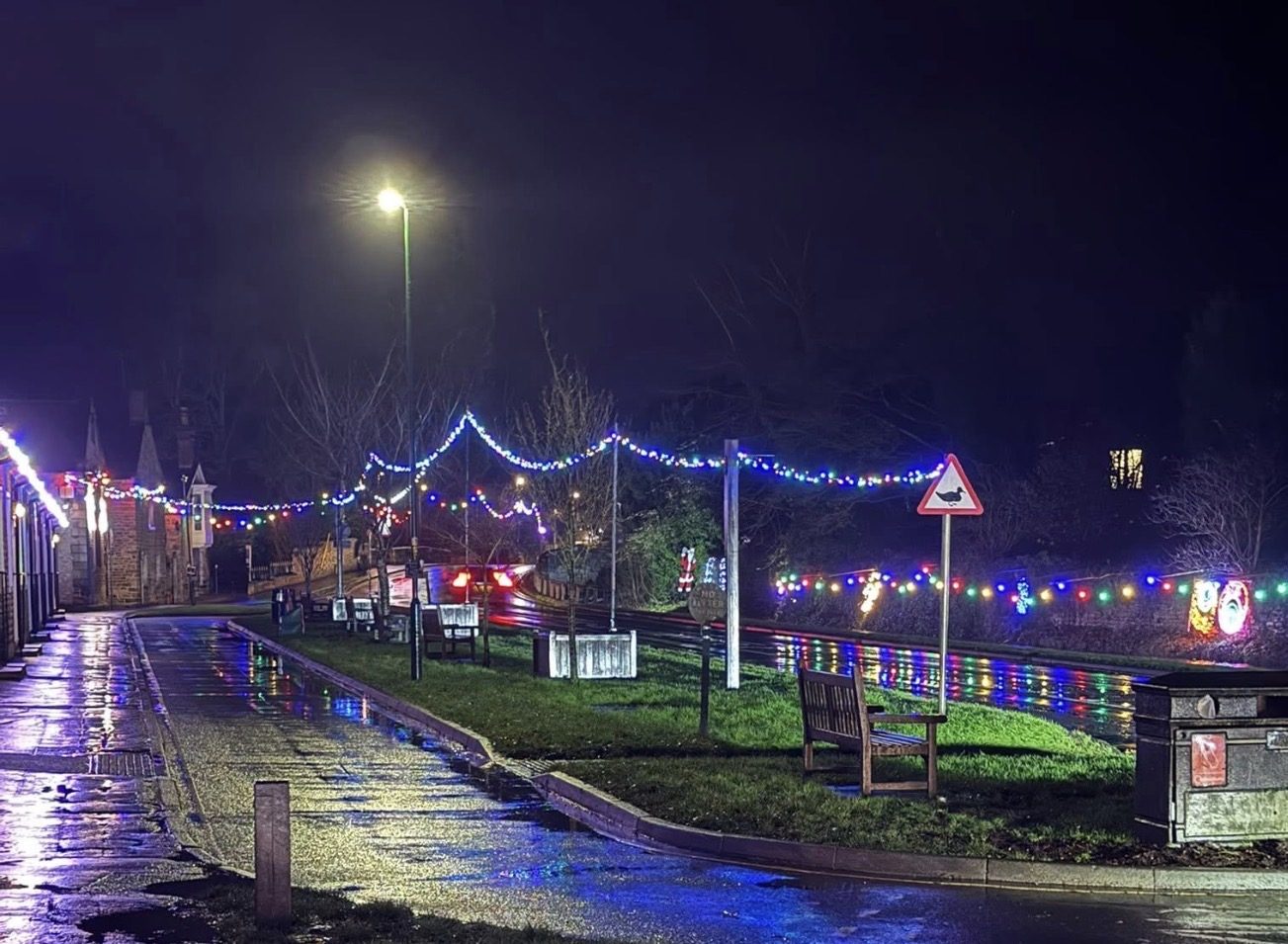 Coloured Christmas lights hang above the road and along Thornton Beck in the centre of Thornton-le-Dale at night, reflecting on the wet road and pavement beside benches.