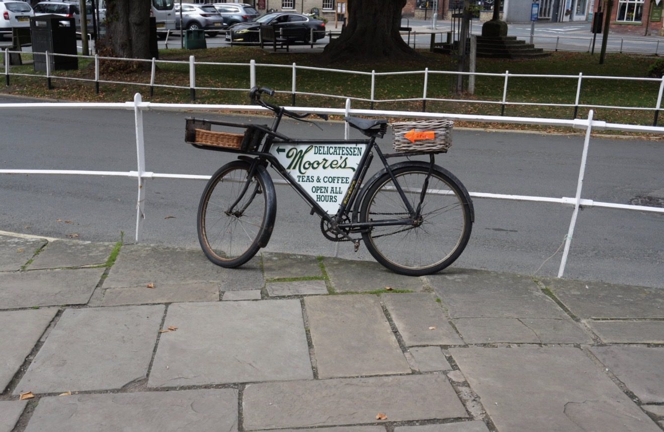 A black delivery bicycle with a Moore's Delicatessen sign, leaning on white railings opposite the green