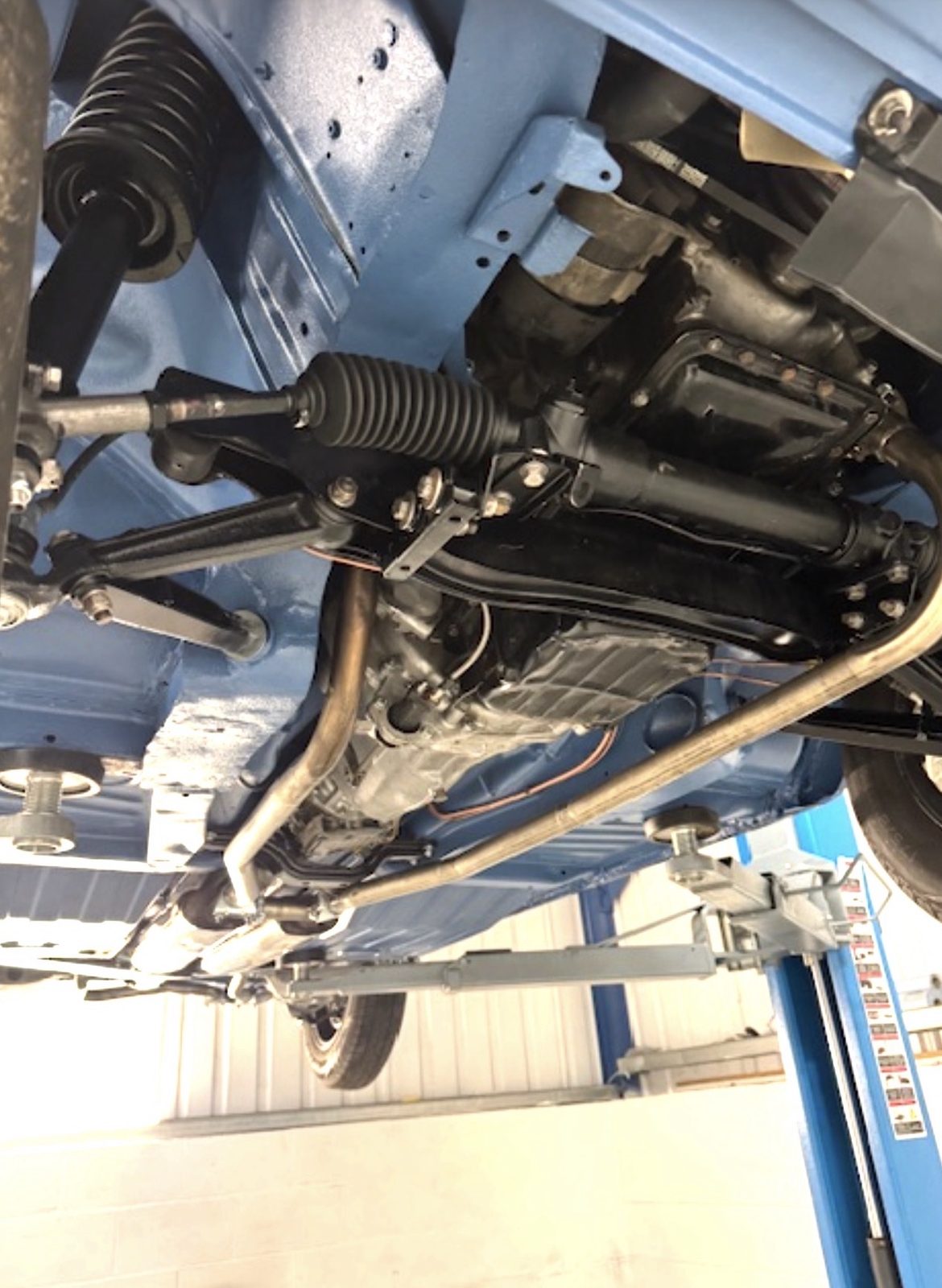 The underside of a Triumph Stag raised on a workshop lift, showing the freshly painted floor, black steering rack and wishbones, a front coil spring and twin exhaust pipes.