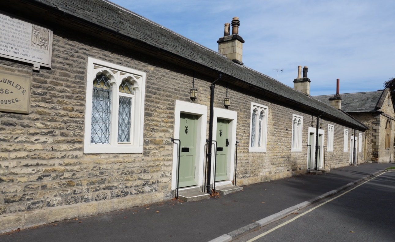 A single-storey stone row of almshouses with green doors and pointed windows, and the founding plaque on the wall