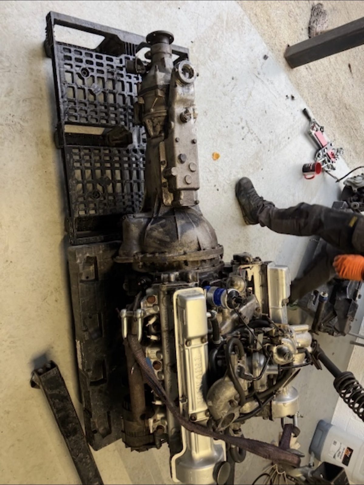 A Triumph V8 engine and gearbox out of the car and resting on a pallet on a workshop floor, with a lifting strap around it and a mechanic's booted feet and gloved hand at the edge.