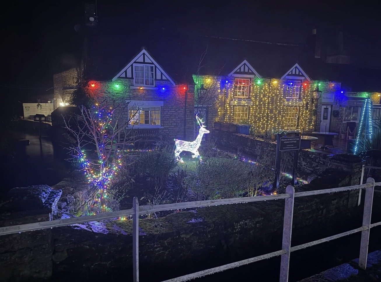 An illuminated reindeer and a tree wrapped in coloured Christmas lights stand beside Thornton Beck in Thornton-le-Dale, with lit stone cottages behind them at night.