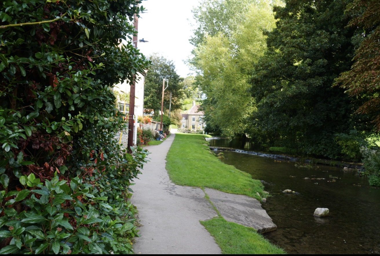 A paved path and grass verge running beside Thornton Beck, with a low weir and a house at the far end