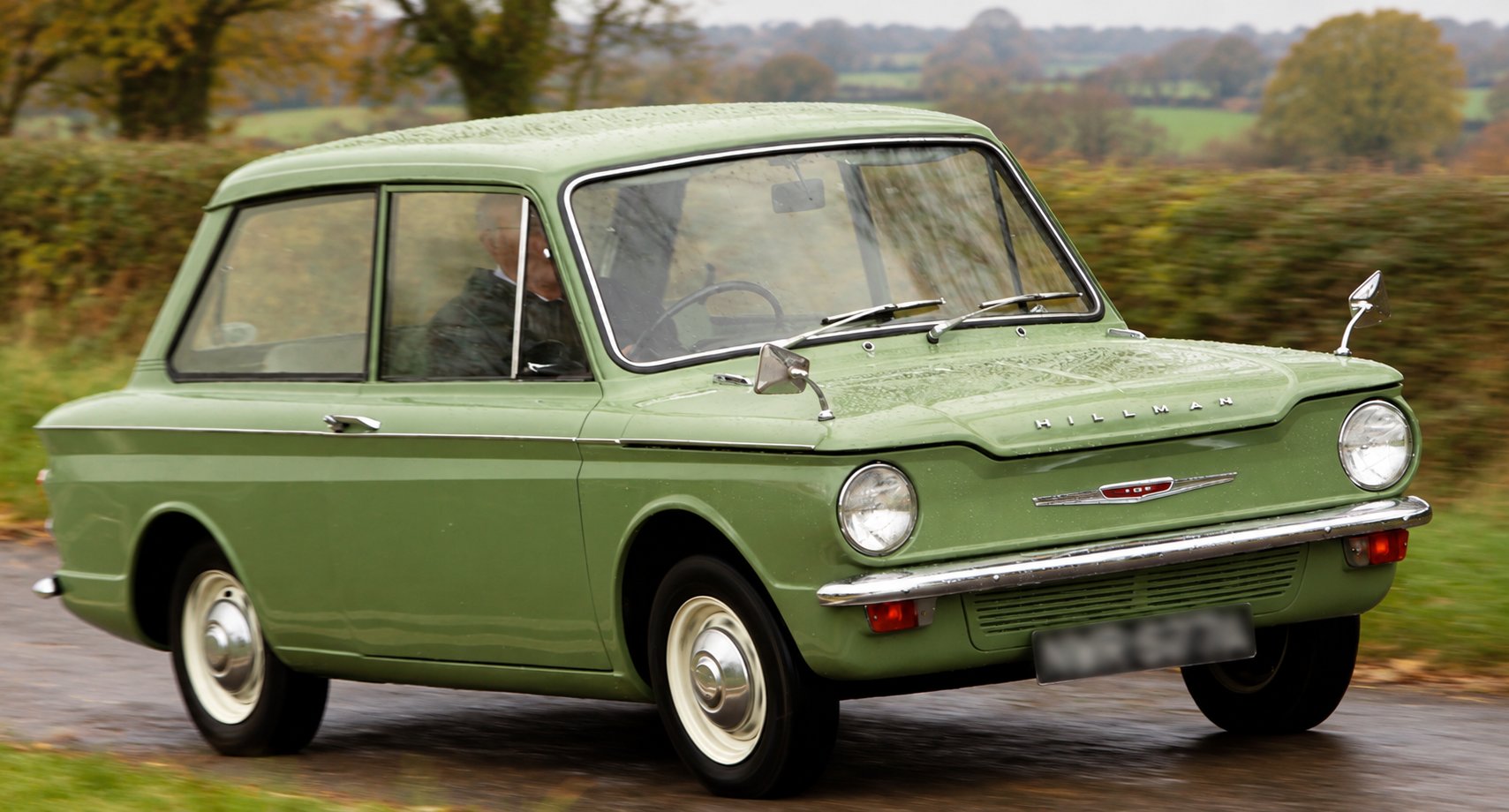 A green Hillman Imp saloon with white wheels and chrome bumpers being driven along a wet country lane, with hedgerows and autumn trees behind