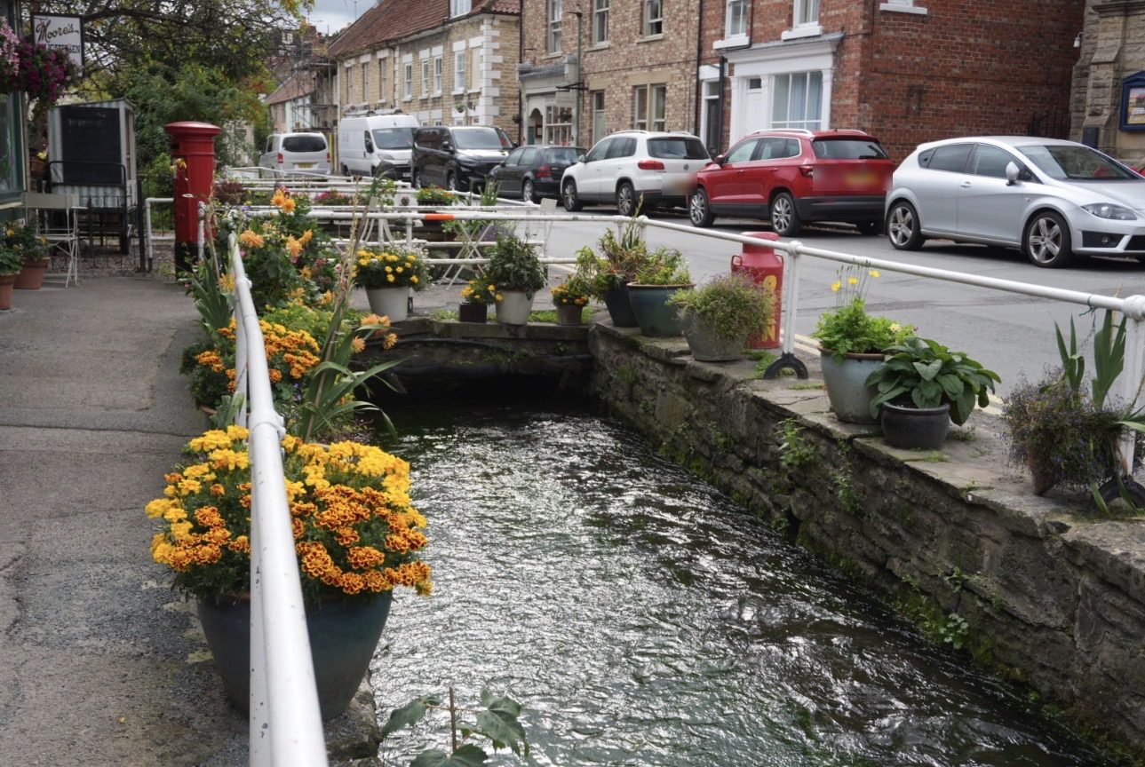 Thornton Beck flowing between a stone wall and white railings lined with pots of marigolds, with parked cars beyond