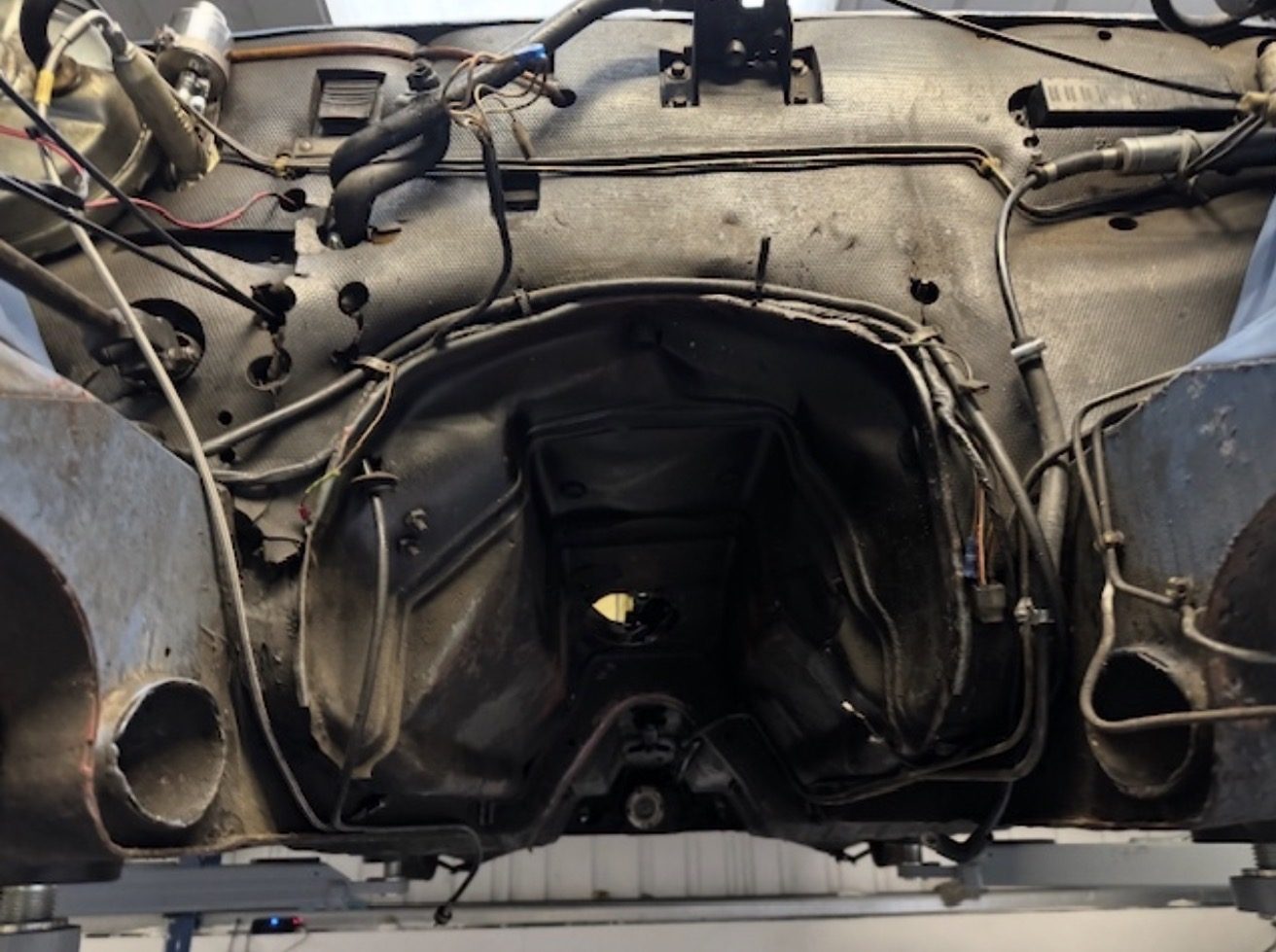 The empty engine bay and bulkhead of a Triumph Stag on a workshop lift, with the engine and gearbox out, showing black insulation on the firewall and exposed wiring, brake and clutch lines.