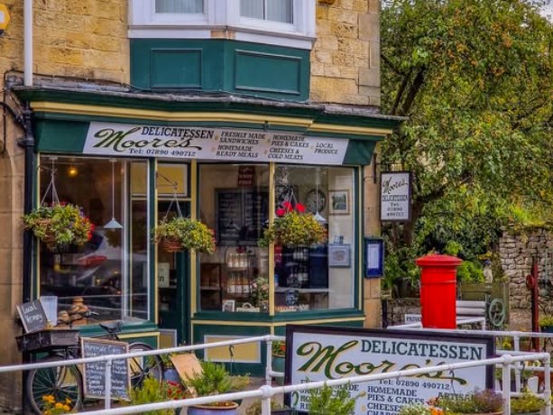 The Moores Delicatessen shopfront on Maltongate in Thornton-le-Dale, with its green fascia sign, hanging flower baskets and a red pillar box outside