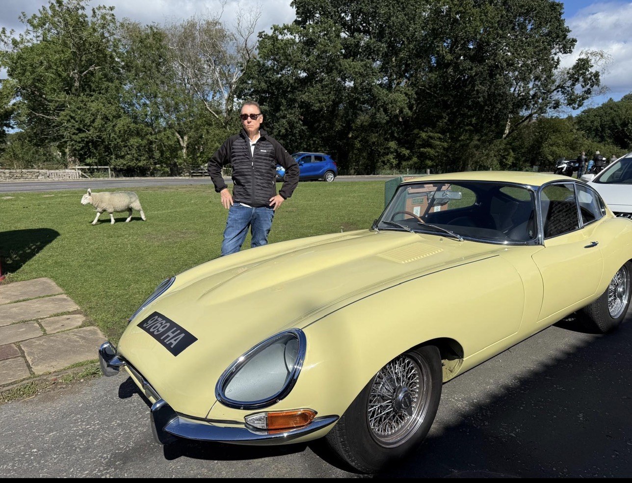 Jon Cosson standing beside his pale yellow Jaguar E-Type, registration 9769 HA, parked by a grass verge, with a sheep grazing behind.