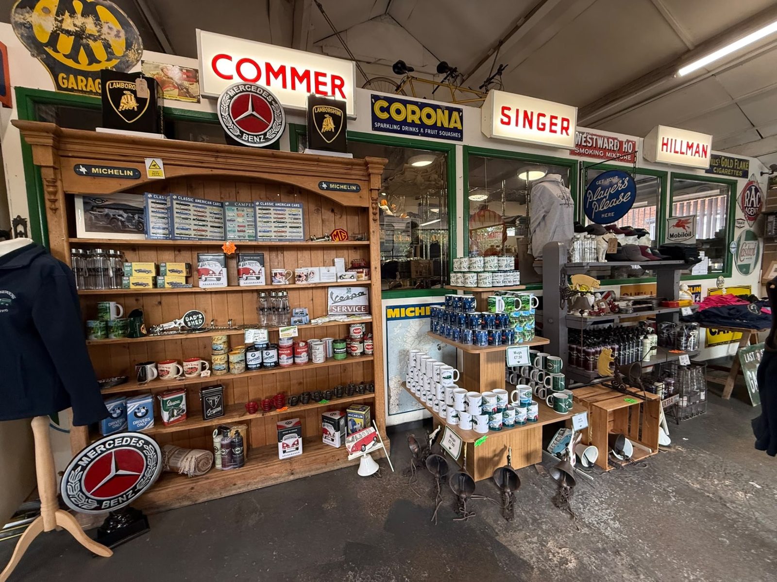Inside the gift shop: a wooden dresser of tins and mugs beneath enamel signs for Commer, Corona, Singer, Hillman and Mercedes-Benz