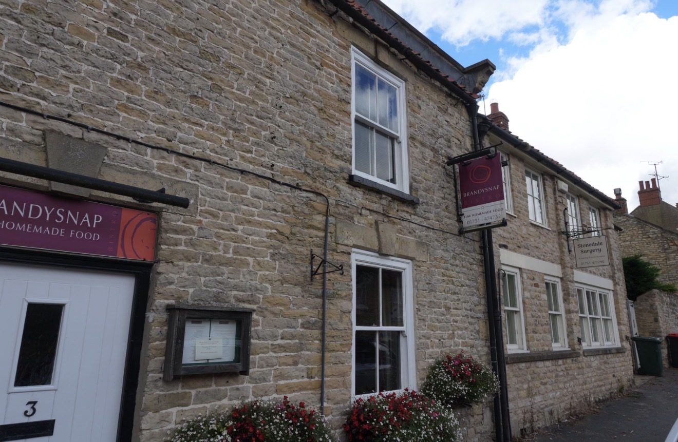 A stone building with the Brandysnap sign and flower baskets, and the Stonedale Surgery sign beyond