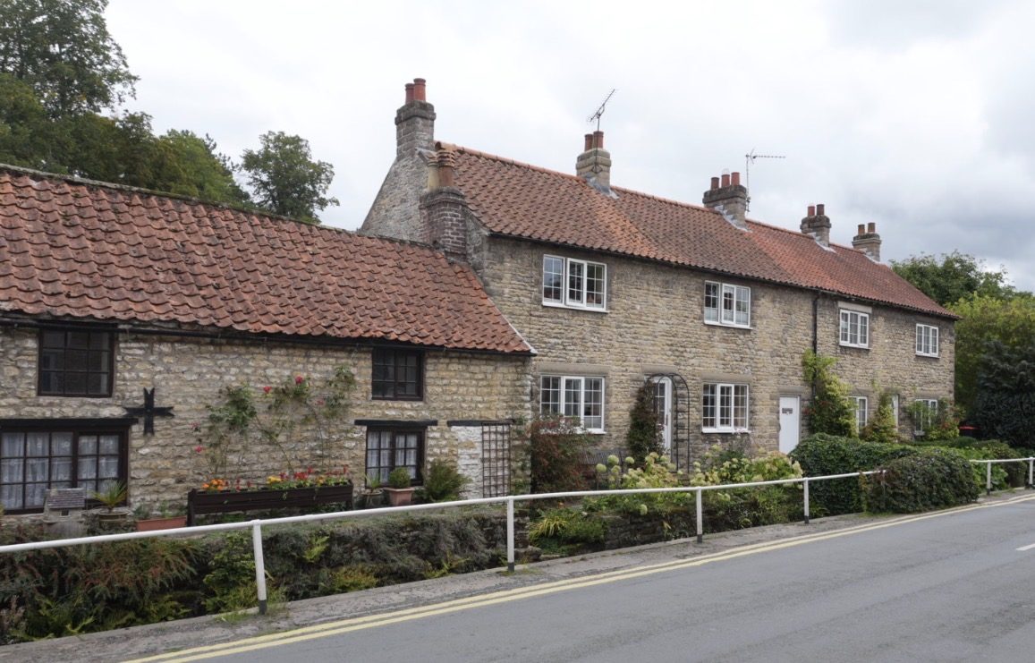 A row of stone cottages with red pantile roofs behind white railings beside the road