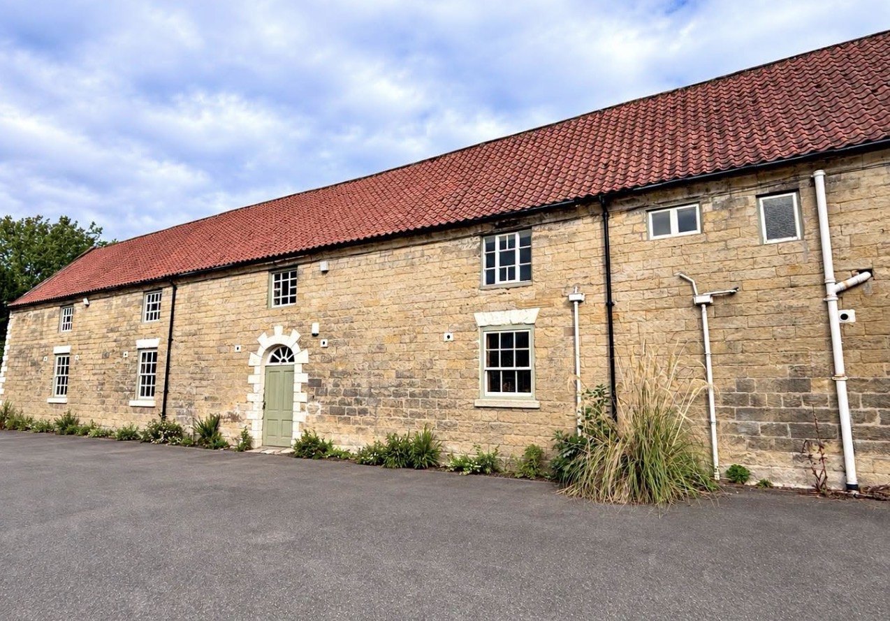 The Old Stables on Church Lane in Thornton-le-Dale: a long buff sandstone stable conversion under a terracotta pantile roof, with an arched doorway and pale green door beneath a fanlight, pale sash windows and a tarmac forecourt in front