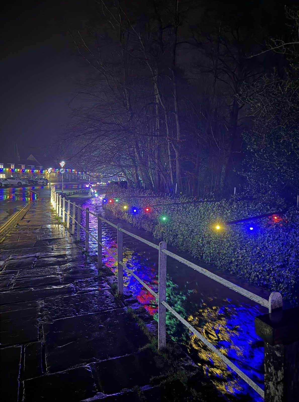 Coloured Christmas lights along Thornton Beck in the centre of Thornton-le-Dale reflect in the water beside the wet footpath and white railings at night.