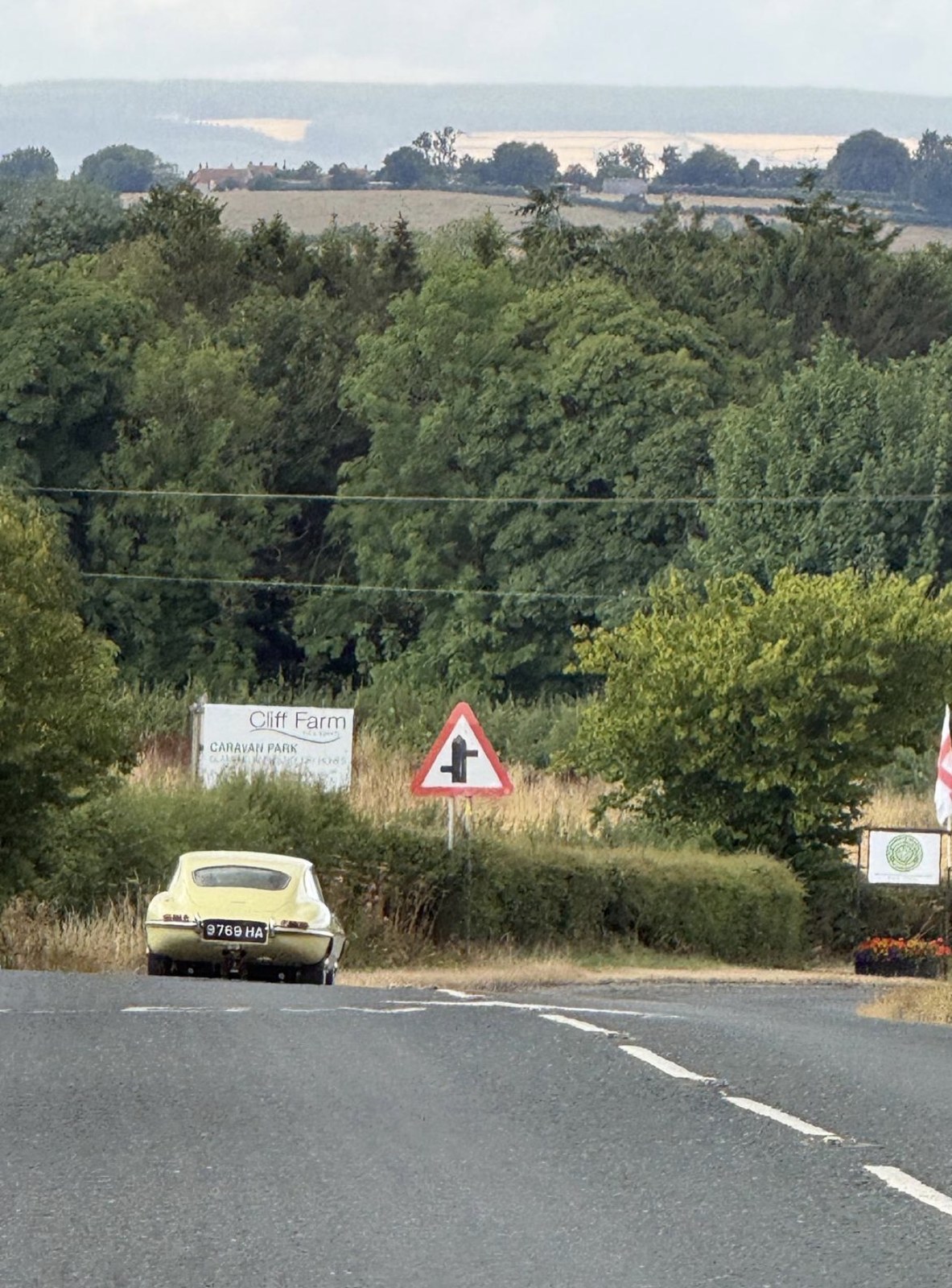 A pale yellow Jaguar E-Type with registration 9769 HA seen from behind on a descending country road, with a junction warning sign, trees and fields ahead.