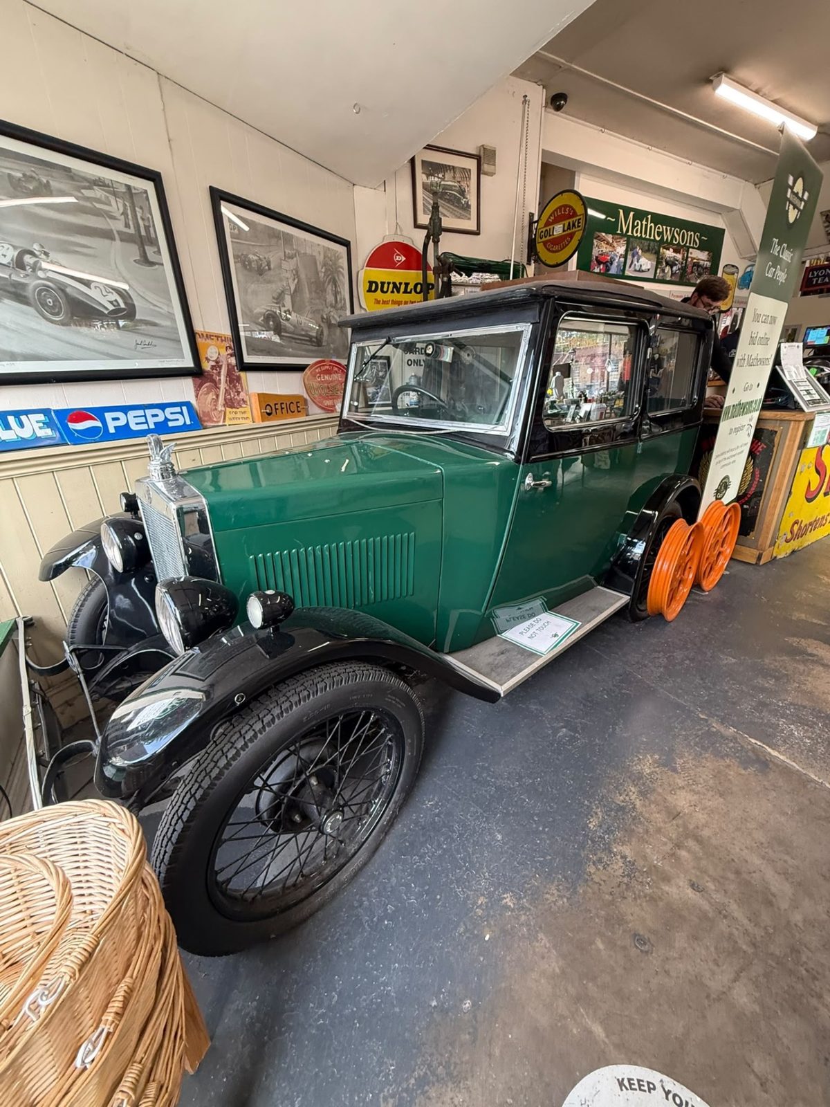 A green 1930s-style saloon with black wings and wire wheels parked indoors among framed motor-racing photographs and enamel signs, including Pepsi and Dunlop