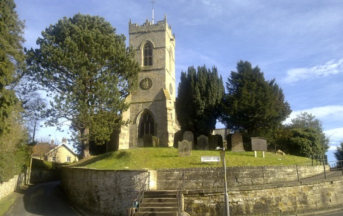 All Saints Church seen from the road below, its battlemented tower rising above a grassy churchyard mound, with a Church Gate signpost and a flight of steps up through the stone retaining wall