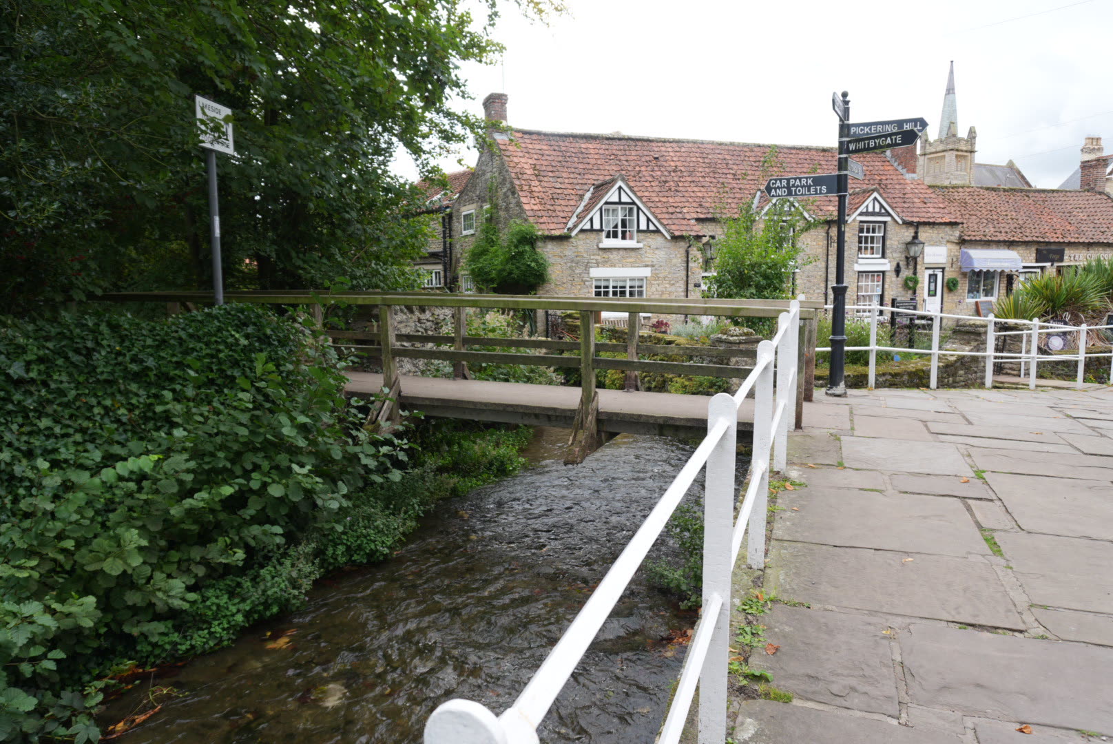 A wooden footbridge over Thornton Beck beside a signpost pointing to Pickering Hill, Whitbygate and the car park and toilets