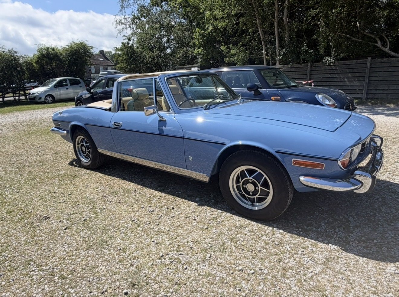 A light blue Triumph Stag with its soft top down parked on a gravel forecourt, seen from the front three-quarter, with other cars and a stone building behind.