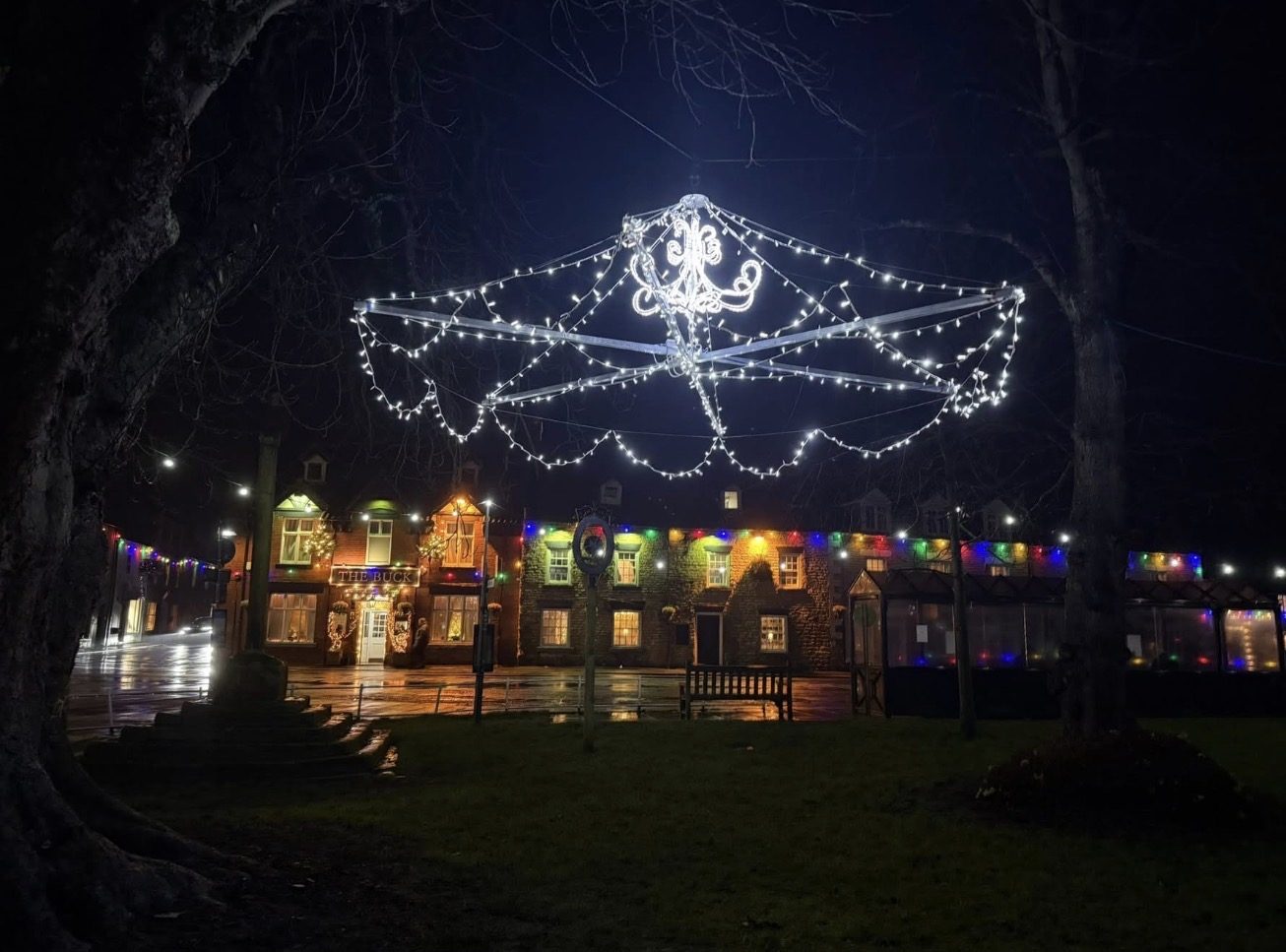 A white chandelier-shaped Christmas light display hangs above the village green in Thornton-le-Dale at night, with The Buck Inn and coloured lights across the road.